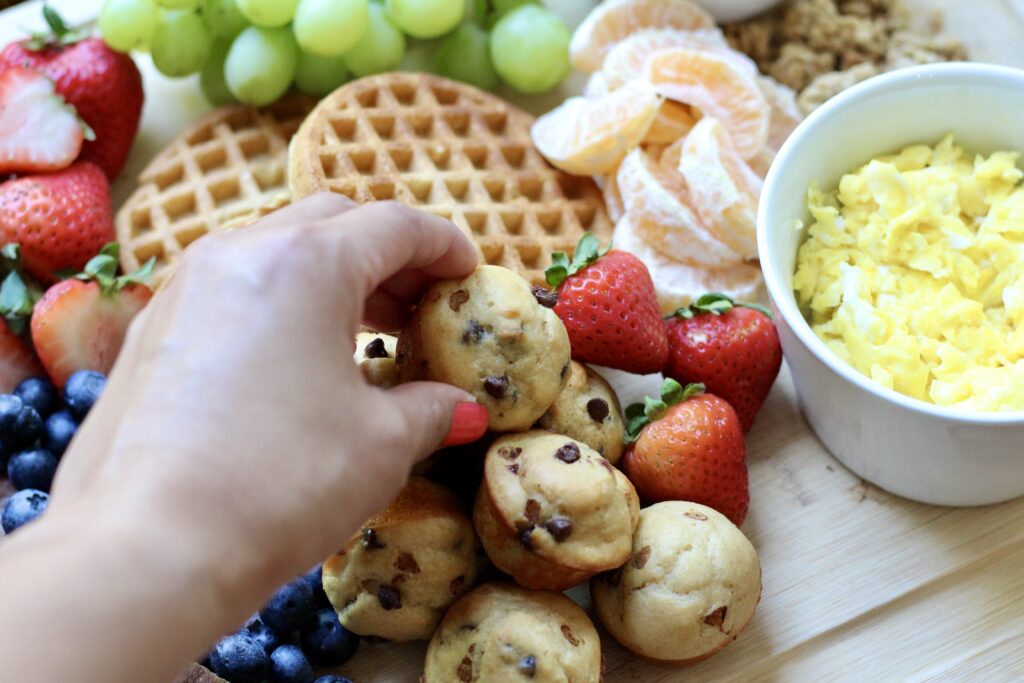 Hand reaching for a mini chocolate chip protein muffin from a breakfast board with waffles, fruit, and eggs