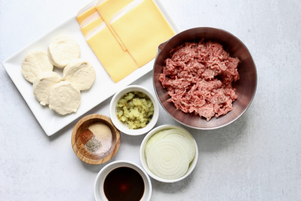 Ingredients for cheeseburger bombs including ground beef, biscuits, American cheese, pickles and onion